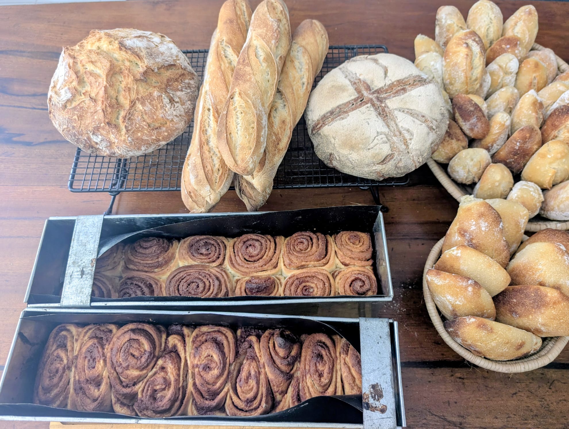 Loaves, baguettes, bread rolls and trays of swirled buns arranged on a wooden table.